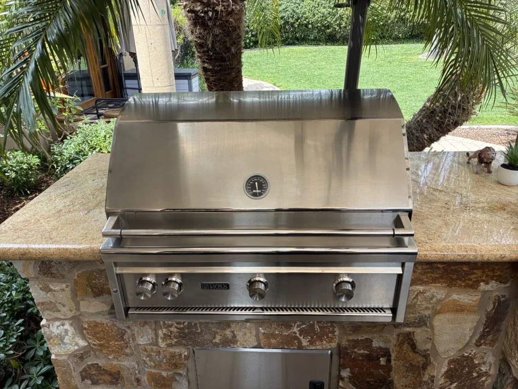 A polished 36-inch Lynx built-in grill in a stone and granite island surrounded by palms